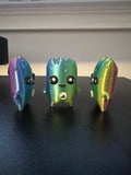May include: Three small, iridescent, 3D-printed figures with smooth, rounded bodies. Each figure features a gradient of blue, green, yellow, and purple colours, large black eyes with white highlights, and a small white circular detail on the torso.