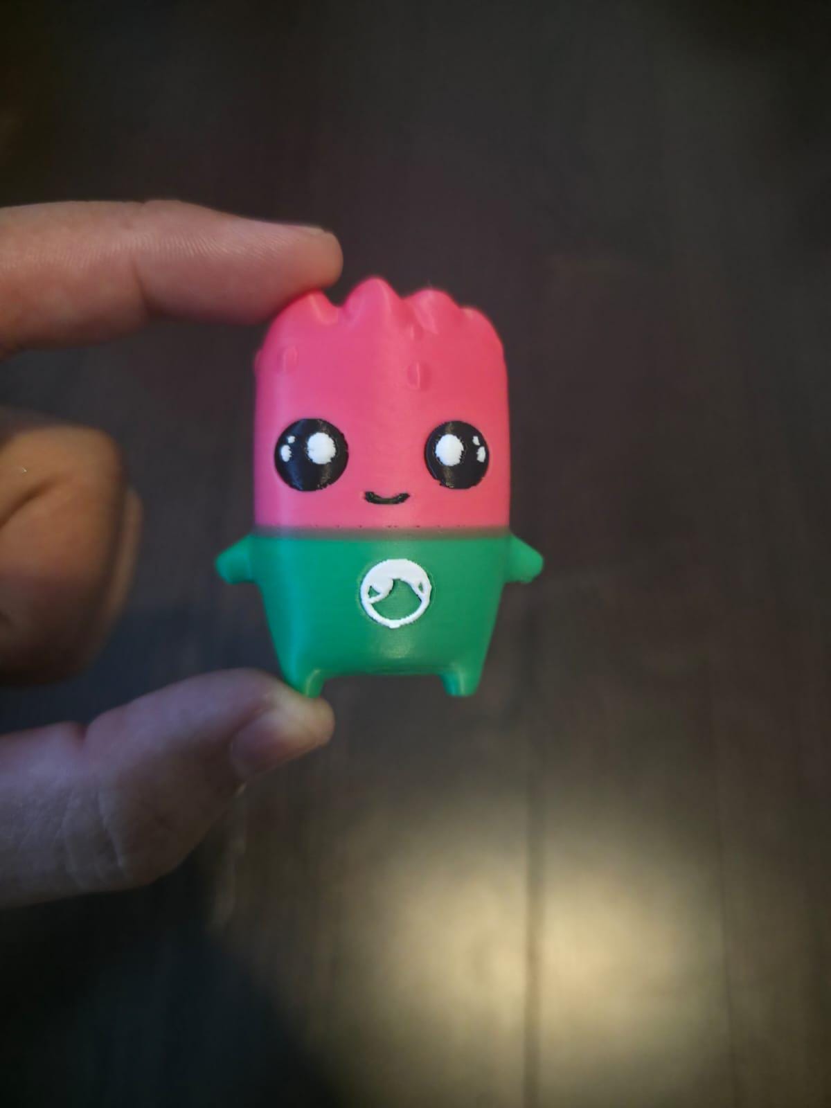 May include: A small, stylised figurine shaped like a watermelon slice with a pink top and a green bottom. It features two large black eyes with white highlights, a simple black smile, and a small white circular logo on the green section.